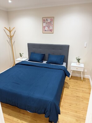 Une chambre à coucher avec un grand lit, une tête de lit rembourrée de couleur grise, deux tables de chevet ornées d’objets décoratifs, un plancher en bois et une œuvre d’art murale aux motifs floraux.