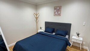 A bedroom with a large bed, a bedside table, a nightstand, a mirror, and a decorative plant.