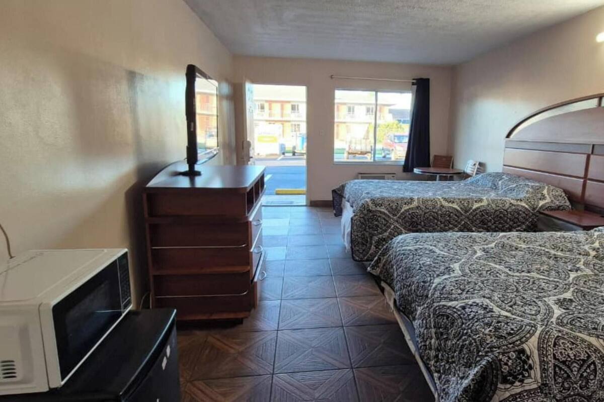 Ein Hotelzimmer mit zwei Betten, einem Schminktisch, einem Fernseher und einem Fenster mit Blick auf Gebäude.