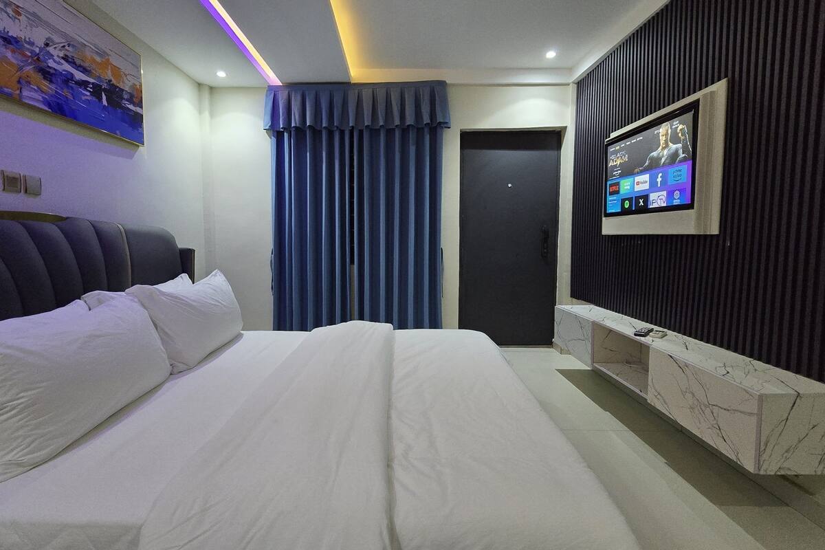 Moderni hotellihuone, jossa on suuri sänky, seinälle asennettu taulutelevisio ja tummanvärinen ovi.