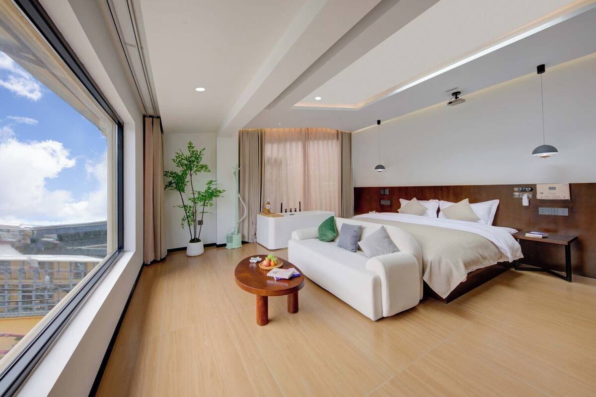 Een moderne hotelkamer met een groot bed, een bank, een bijzettafeltje en uitzicht op de stad door grote ramen.