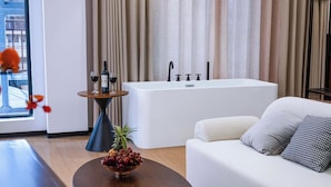 Une chambre d’hôtel moderne avec une baignoire blanche, un canapé blanc et une table ronde en bois sur laquelle est posé un panier de fruits.