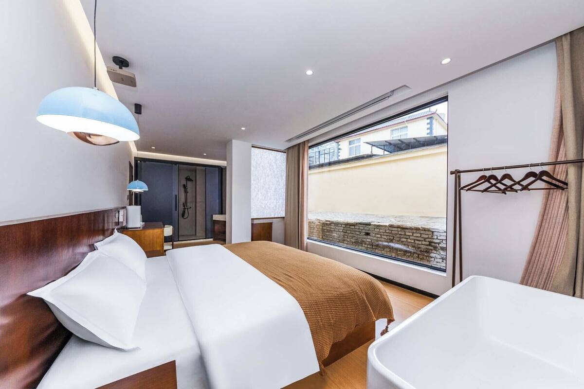 Een moderne hotelkamer met een groot bed, een raam met uitzicht op een bakstenen muur, een badkamer met een wit wastafelmeubel en een hangende lamp.