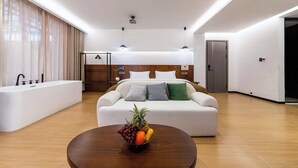 Une chambre d’hôtel moderne avec un canapé blanc, une table ronde en bois sur laquelle sont disposés des fruits, une grande fenêtre avec des rideaux, une baignoire intégrée et un lit avec une tête de lit.