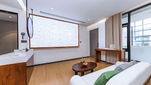 Une chambre d’hôtel moderne dotée d’une grande fenêtre, d’un plancher en bois, d’un canapé blanc, d’une petite table ronde avec des fruits, d’un bureau en bois et d’un écran de projection.