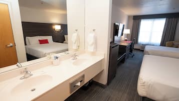 A hotel room with a large mirror, two beds, a desk with a chair, a TV, and a bathroom with a double sink.