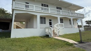 Um edifício branco de dois andares com varanda, alpendre coberto e escadaria que leva ao andar superior.