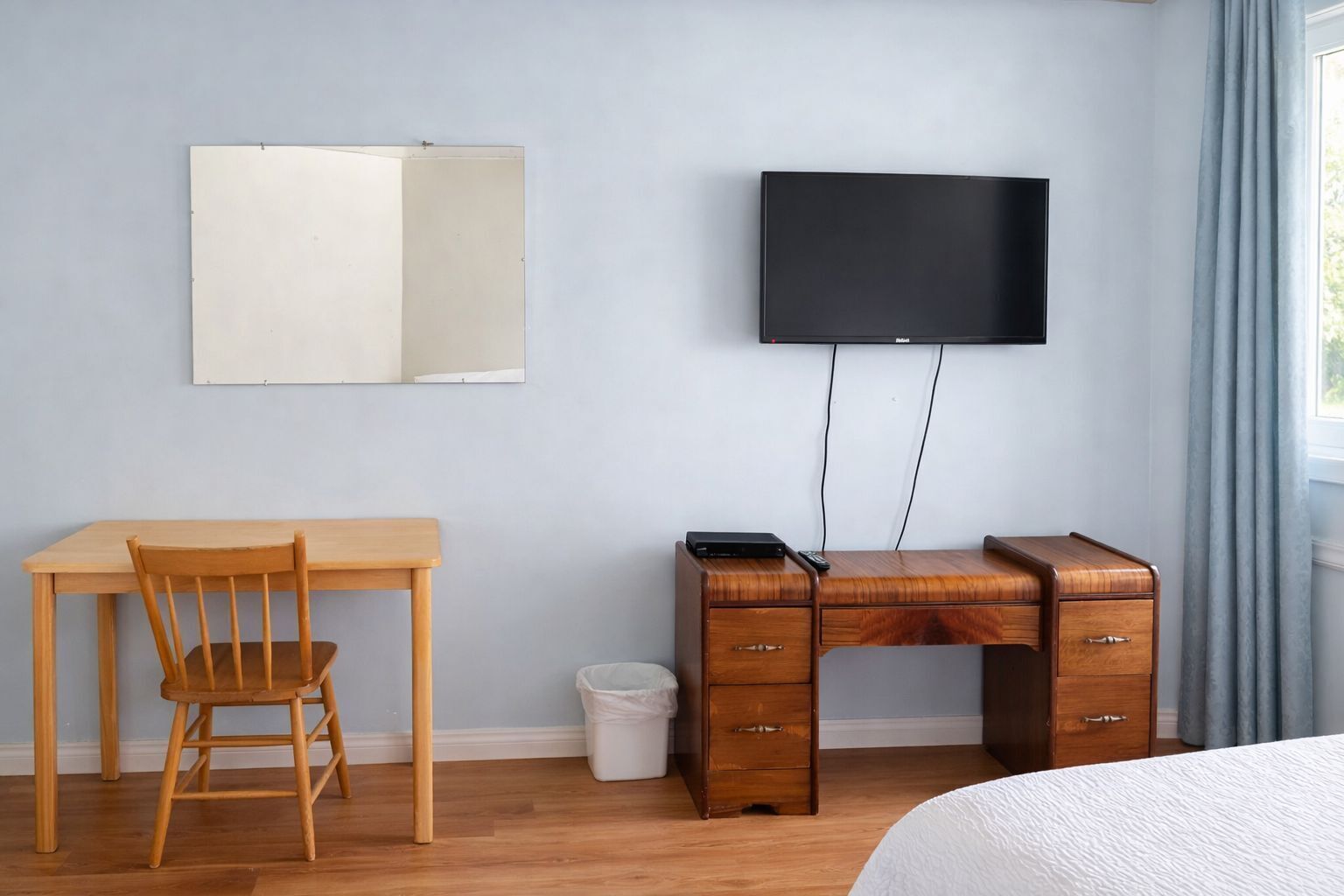 Un dormitorio con cama, un escritorio de madera con cajones, una silla, televisor en la pared y una ventana con cortinas.
