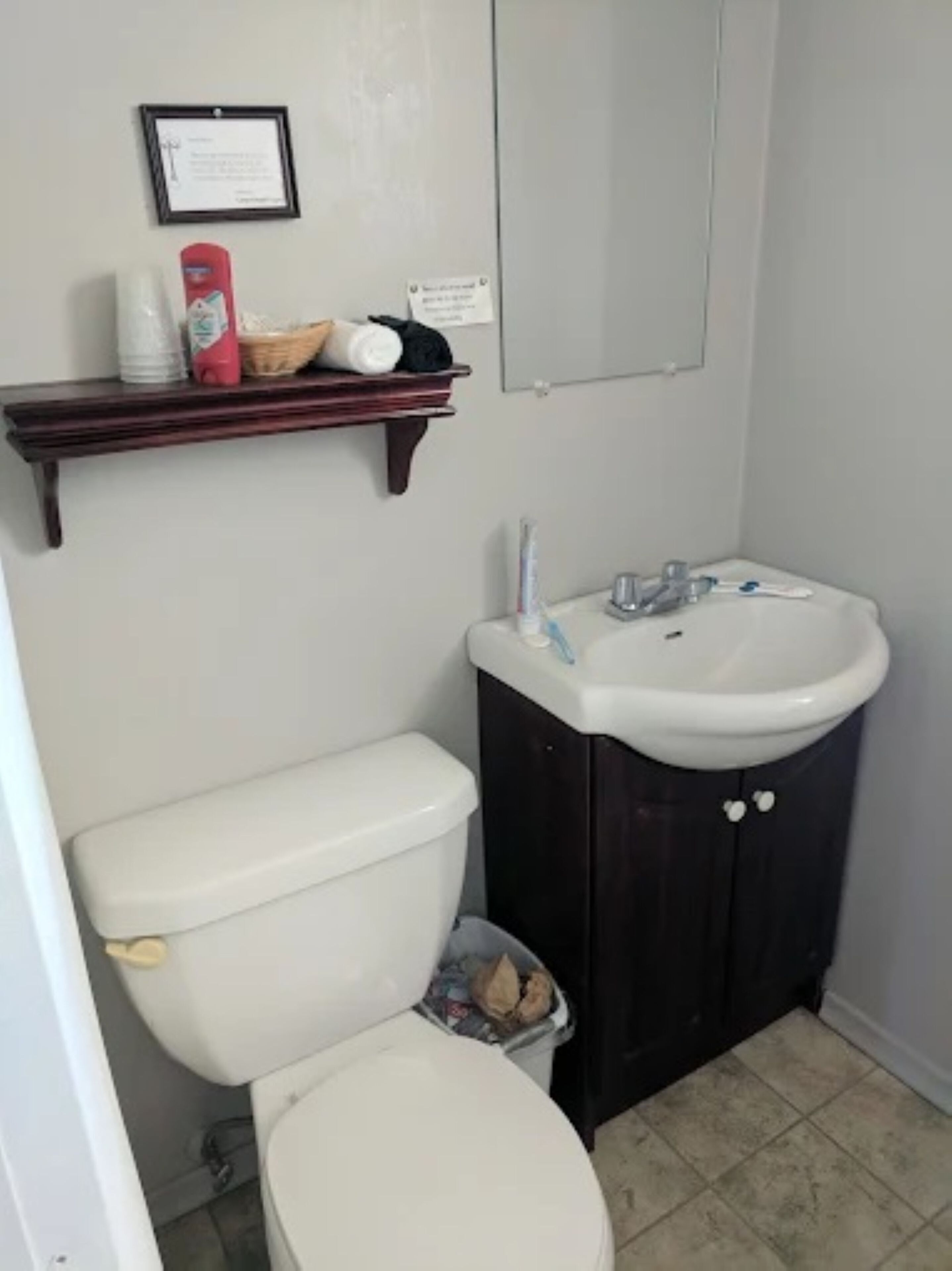 Een kleine badkamer met een wit toilet, een donker houten kastje met een wastafel, en een plank boven het toilet met toiletartikelen.