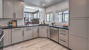 Une cuisine moderne avec des meubles de couleur gris clair, un plan de travail noir et un lave-vaisselle en acier inoxydable. On y trouve une fenêtre avec des stores, un évier et une petite plante posée sur le plan de travail.