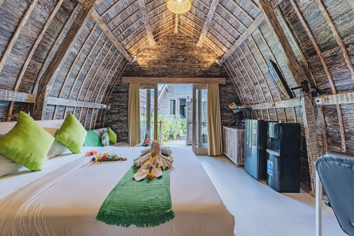 Ein gemütliches, rustikales Schlafzimmer mit hohen Holzbalken, einem großen Bett mit grünen Kissen und Blick ins Freie.