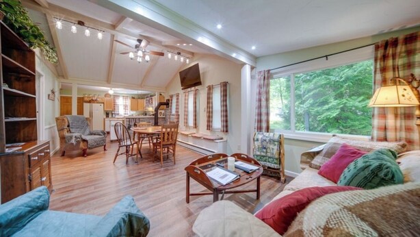 A cozy living room with a wooden floor, a dining area, and a kitchen in the background. There is a large window with curtains, a ceiling fan, and a lamp on the side table. - Earth's River Retreat: Kayaks, Wineries and GOTL (Austinburg Township)