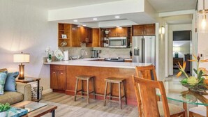 A modern kitchen with wooden cabinets, a stainless steel refrigerator, and a large island with stools. - Beachfront • Pool + Hot Tub • Pickleball, Tennis (Lahaina)