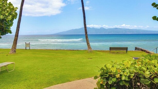 A grassy area with a bench and a sign, overlooking a beach with clear blue water and a mountain in the distance. - Beachfront • Pool + Hot Tub • Pickleball, Tennis (Lahaina)