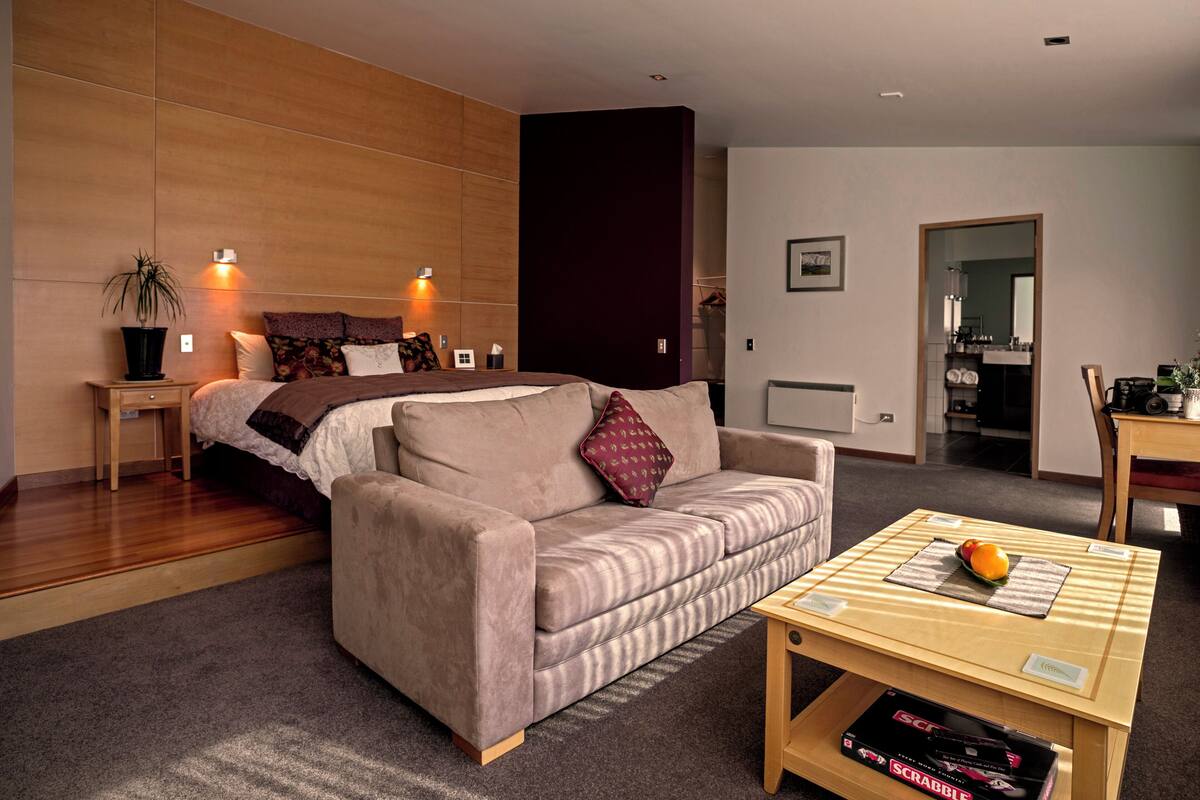 A modern living room with a sofa, a coffee table with a fruit bowl, and a bed with a headboard.