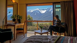A room with a large window offering a mountain view, a desk with a lamp, a chair, and a bed.