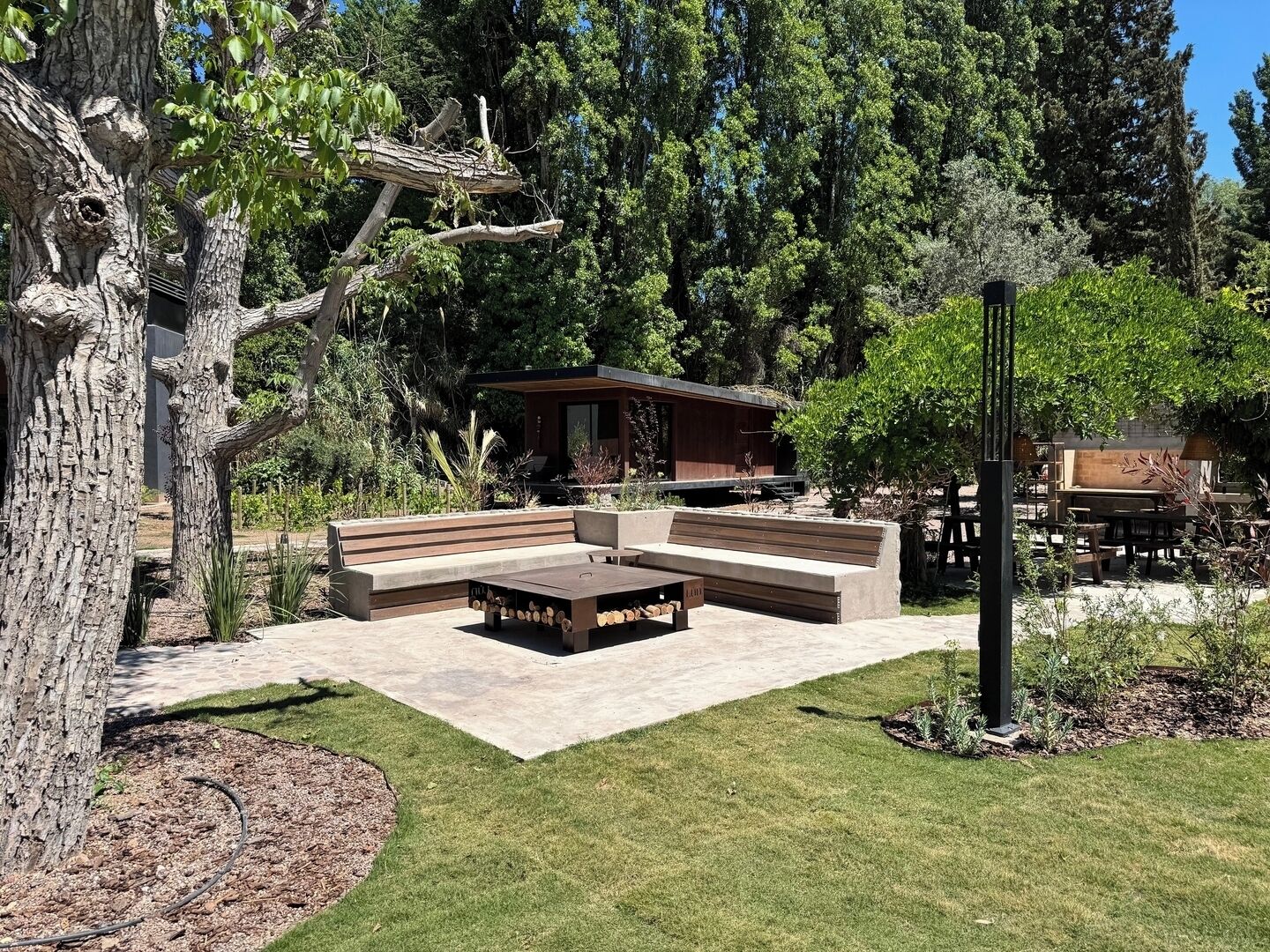 An outdoor seating area with a built-in bench and table, surrounded by trees and a small building.