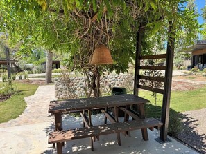 Een houten picknicktafel onder een pergola, met een geweven mand eraan, omringd door een stenen muur en groen.