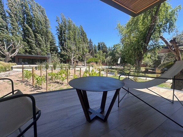 Een terras buiten met een ronde tafel en twee stoelen, omgeven door een houten terras en een uitzicht op een aangelegde tuin met bomen en een gebouw op de achtergrond.