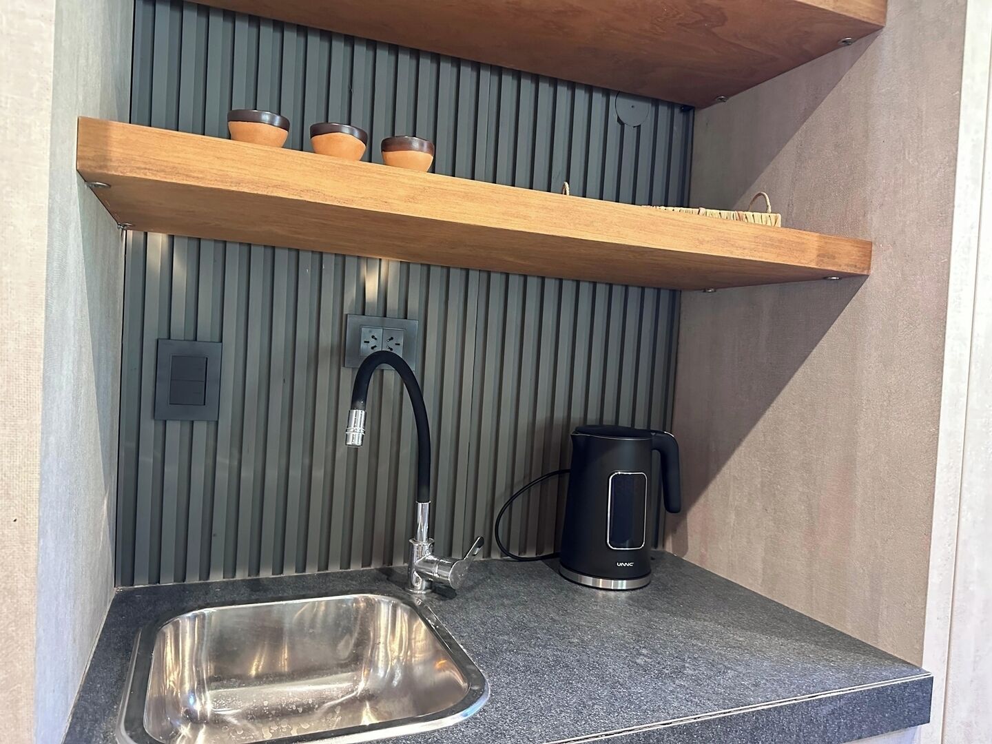 Una cocina compacta con un lavamanos de acero inoxidable, una tetera eléctrica negra y un estante de madera sobre un enchufe.