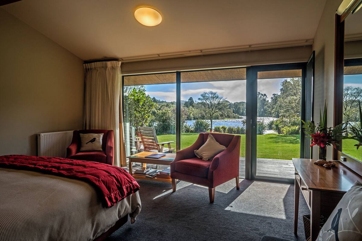 A hotel room with a large bed, a red armchair, a wooden table, and a view of a lush garden and lake through large glass doors.
