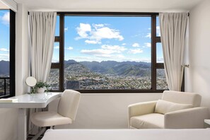 Ein Hotelzimmer mit großem Fenster, Schreibtisch, zwei Stühlen und Blick auf Stadt und Berge.