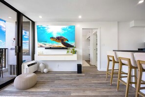 Ein modernes Wohnzimmer mit einem großen Flachbildfernseher an der Wand, Holzboden, einem runden Hocker und einem Essbereich mit Holzstühlen.
