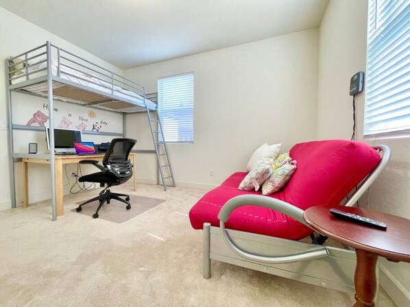 A room with a bunk bed, a desk with a laptop, a red chair, and a small table with a remote control.