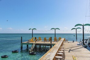A wooden pier extending into clear blue water with yellow chairs and tables, and string lights. - Reef Haven Penthouse | 1BR Sea View Suite (San Pedro)