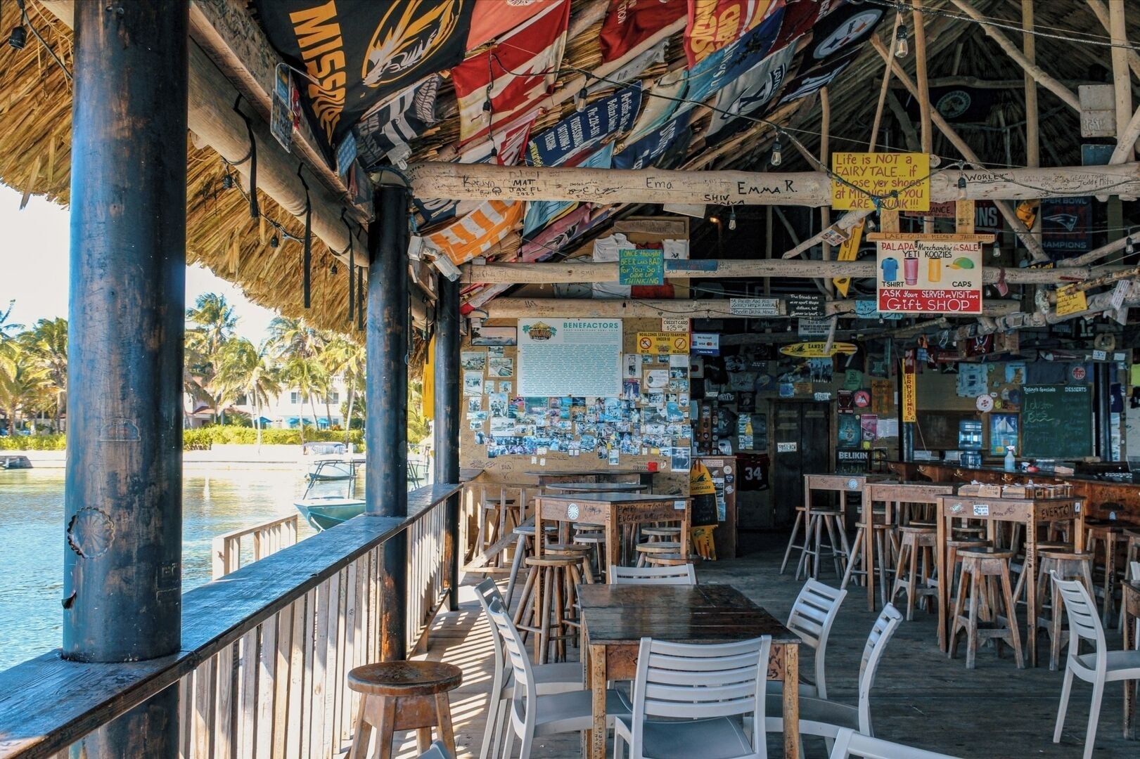 A beachside restaurant with outdoor seating, a thatched roof, and various signs and posters on the walls.