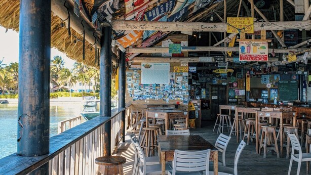 En restaurang vid stranden med uteservering, ett halmtak och diverse skyltar och affischer på väggarna.