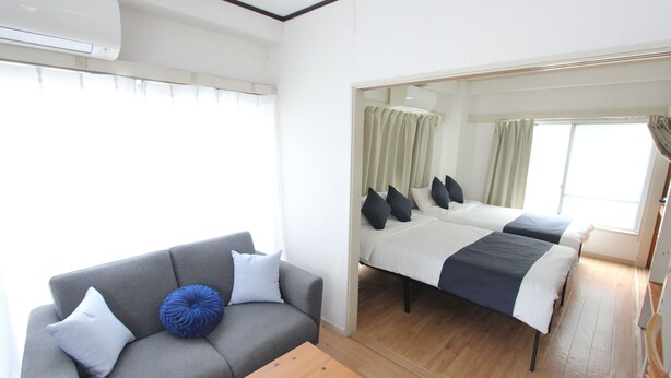 Een kamer met een grijze bank, een houten salontafel, een bed met twee kussens en een raam met gordijnen.