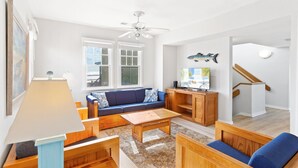 Ein Wohnzimmer mit Sofa, Couchtisch und TV-Möbel. Fenster mit Blick auf den Strand, ein Deckenventilator und eine Treppe sind ebenfalls vorhanden.