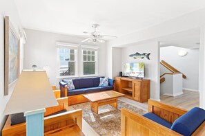 A living room with a sofa, coffee table, and TV stand. There are windows with a view of the beach, a ceiling fan, and a staircase. - Dolphin: Dog-Friendly Buxton Beach Retreat (Buxton)