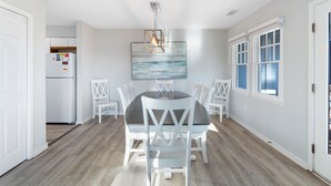 Un coin repas avec une table ronde, des chaises blanches, un lustre moderne et un grand tableau représentant un paysage marin.