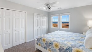 Une chambre à coucher avec un lit, deux fenêtres donnant sur la plage, un ventilateur de plafond et des portes blanches.