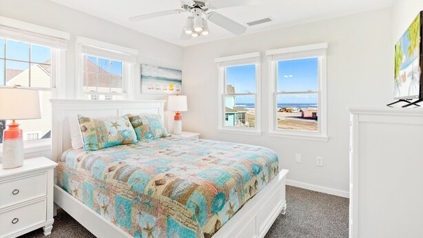 A bedroom with a bed, two nightstands, a ceiling fan, and a view of the beach through large windows.