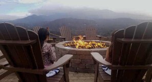 Two wooden Adirondack chairs facing a stone fire pit with flames, set on a patio overlooking a mountainous landscape. - 4th July Westgate Smoky Mtn with heated indoor water park free (Gatlinburg)