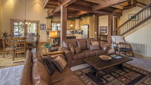 Un ampio soggiorno con un grande divano in pelle marrone, un tavolino in legno e un tappeto decorato. C'è una zona pranzo con sedie di legno e un tavolo, e una cucina sullo sfondo.