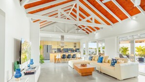 Una sala de estar moderna con un sofá blanco, una mesa de centro y un televisor a la izquierda. En el fondo, se ve una cocina con armarios de madera y un comedor con sillas.