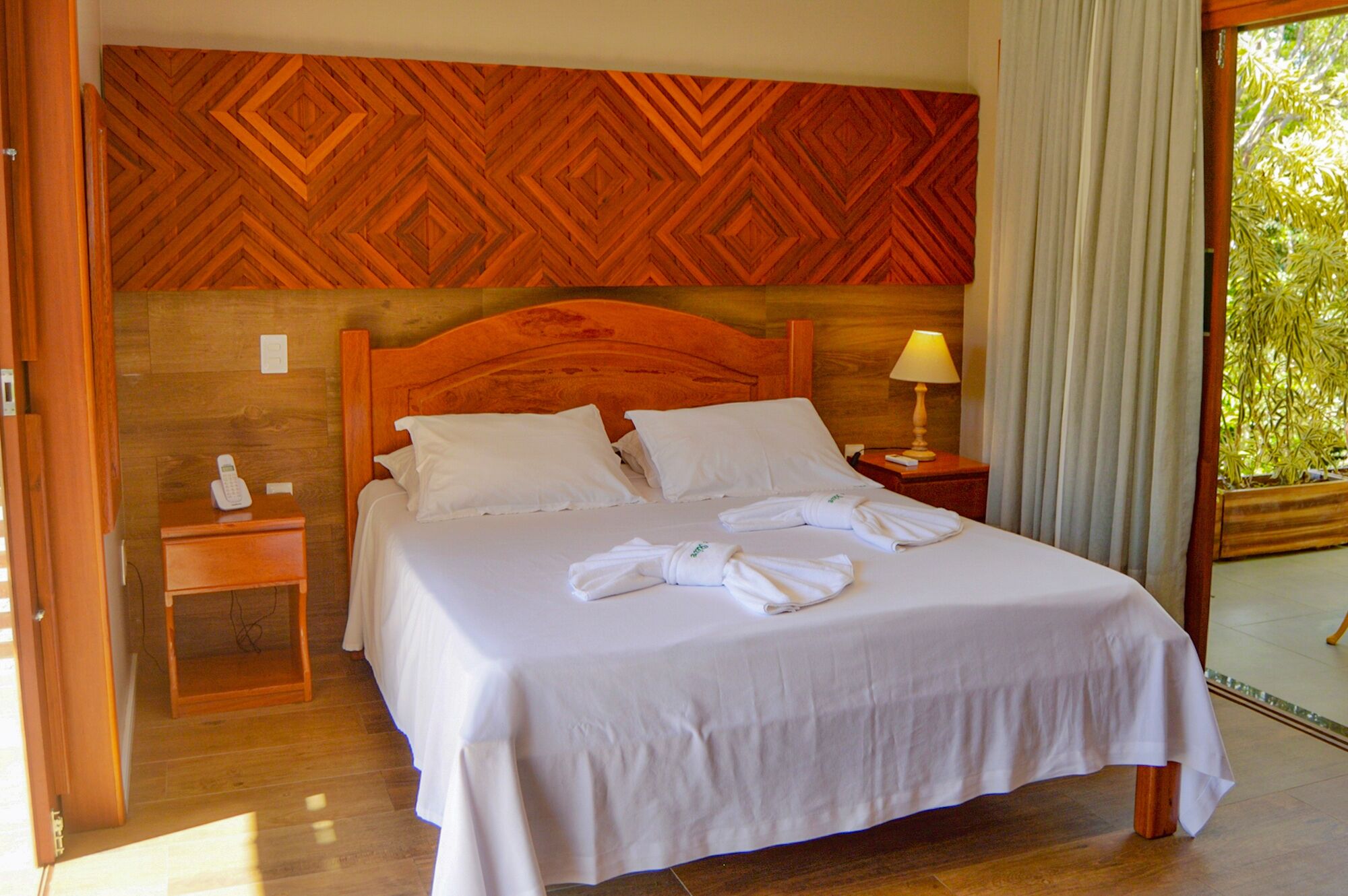 A hotel room with a wooden headboard, a bed with white linens, a bedside table with a lamp, and a view of greenery outside.