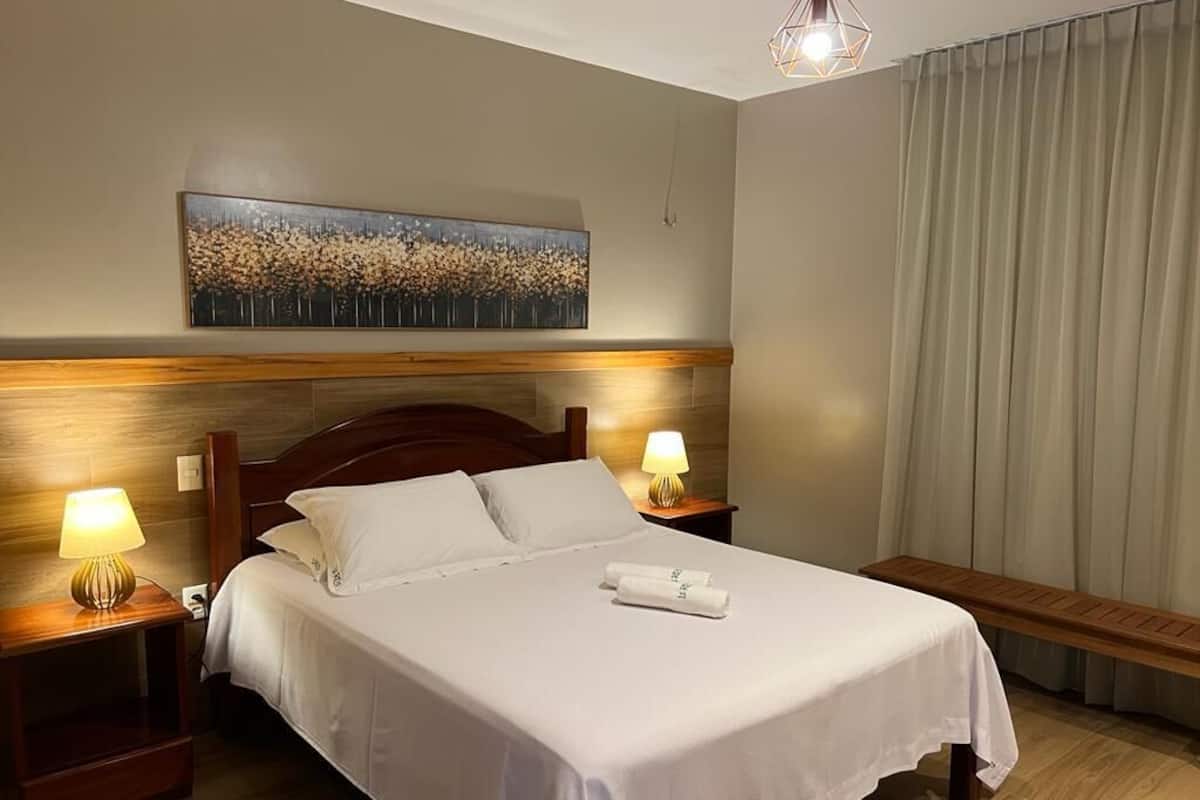 A hotel room with a bed, two bedside lamps, a painting on the wall, a bench, and a large window with curtains.