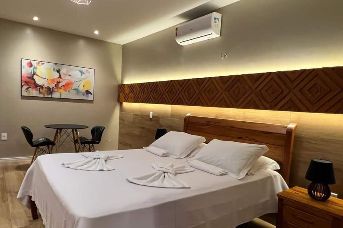 A hotel room with a bed, a desk, a chair, an air conditioner, and a painting on the wall.