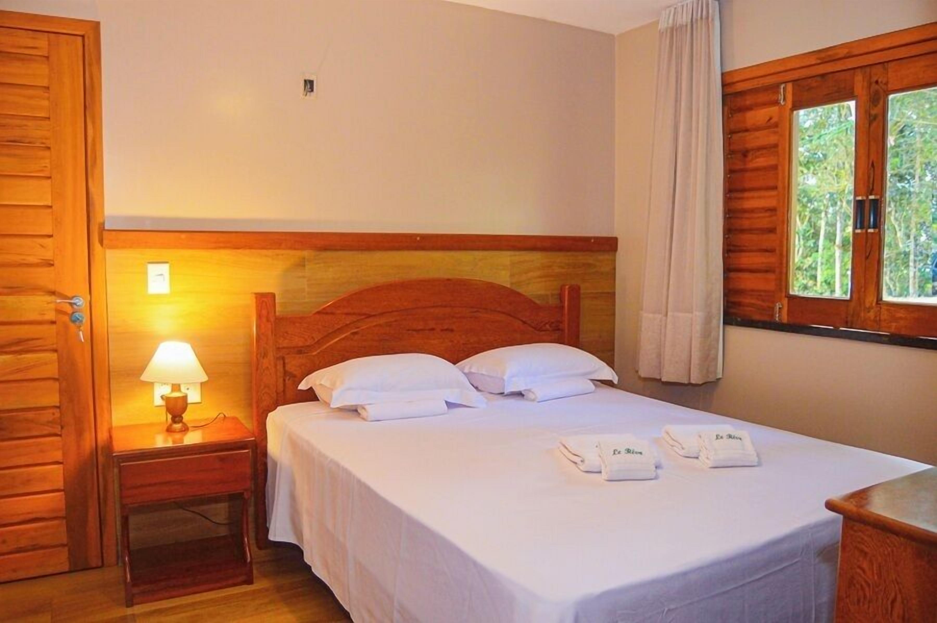 Una cama con estructura de madera, ropa de cama blanca, una lámpara de noche y una ventana con cortinas.