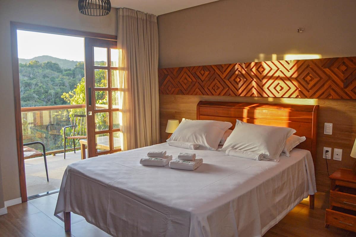 A hotel room with a bed, a wooden headboard, a balcony with a view of the outdoors, and a table with towels.