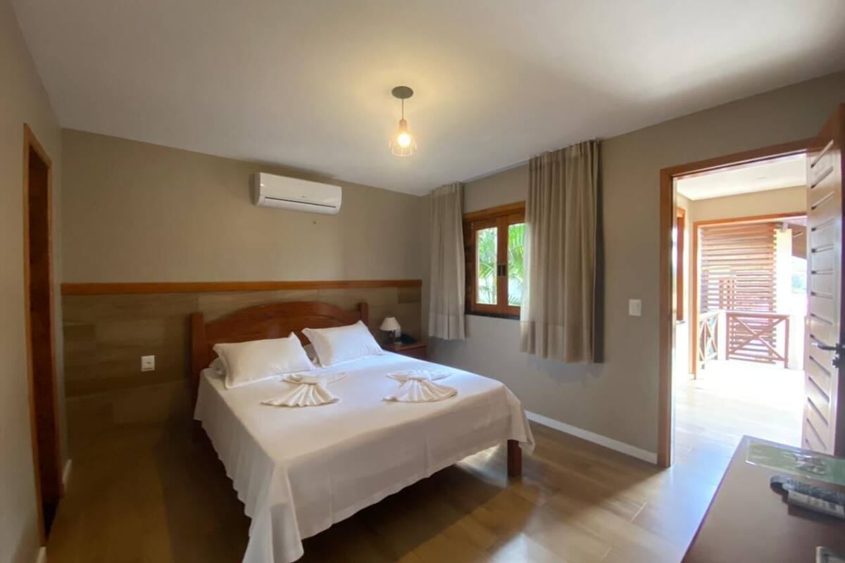 A bedroom with a wooden bed, a nightstand, an air conditioning unit, and a view of the outside through a window.