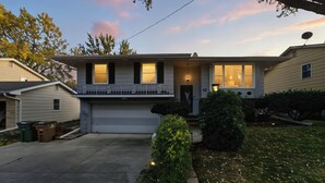 A single-story house with a front porch, a garage, and a small front yard with a tree and a bush. - Cozy Heights Retreat | Baker's Kitchen & Sleeps 10 (Windsor Heights)