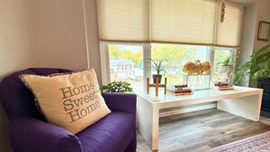A living room with a purple armchair, a white coffee table with books and a plant, and a window with blinds. - Cozy Heights Retreat | Baker's Kitchen & Sleeps 10 (Windsor Heights)
