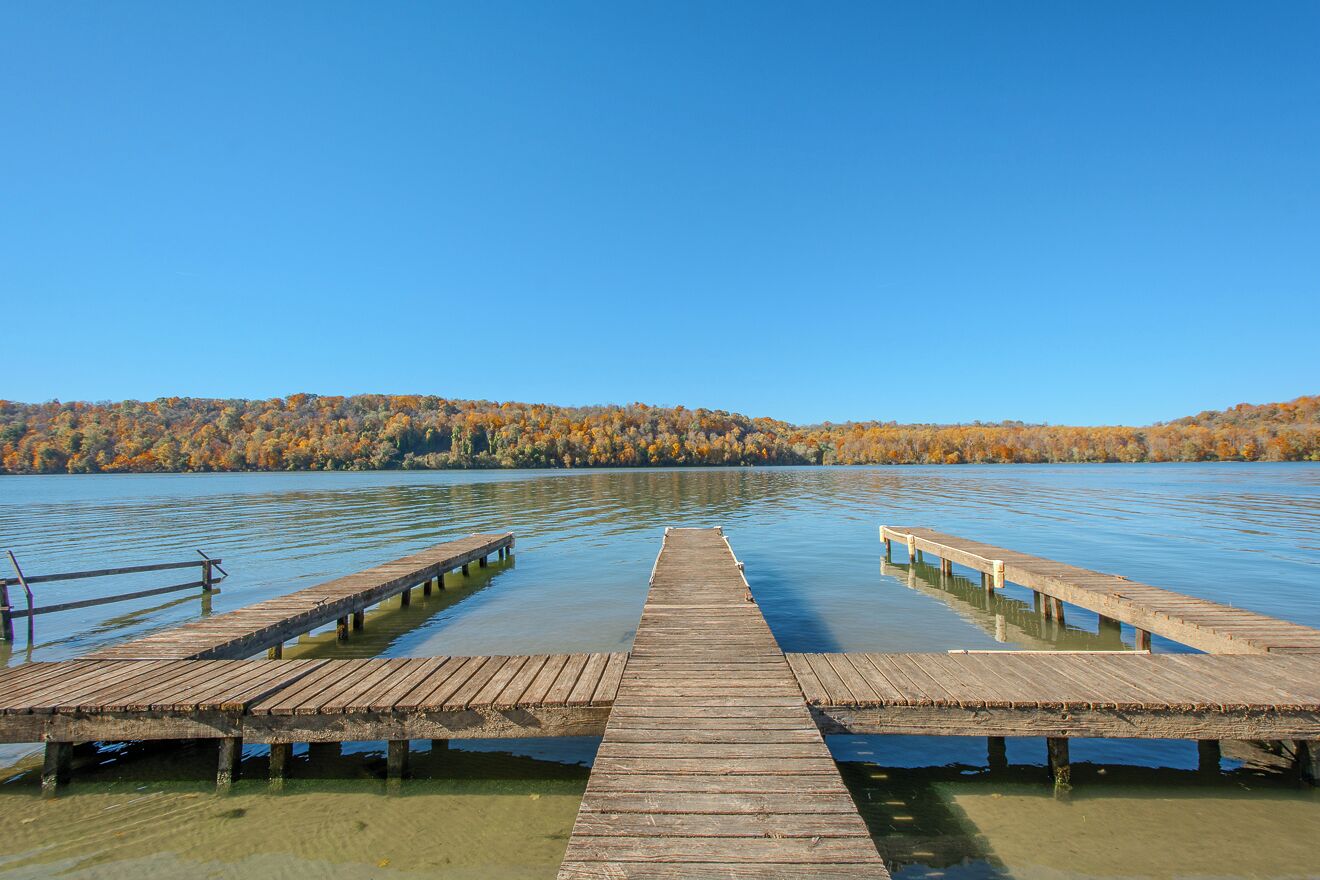 Un muelle de madera que se adentra en un lago tranquilo, con árboles de colores otoñales al fondo.