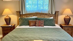 A bedroom with a wooden bed frame, a green quilted bedspread, two bedside lamps, a ceiling fan, and a window with sheer curtains.
